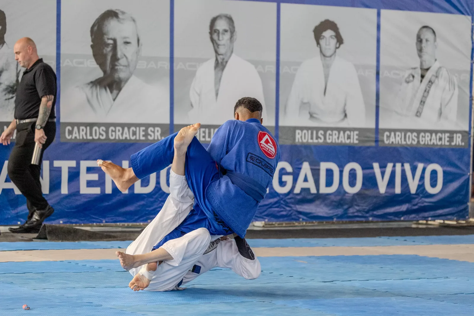 Atletas em queda durante a disputa, diante do painel da Gracie Barra