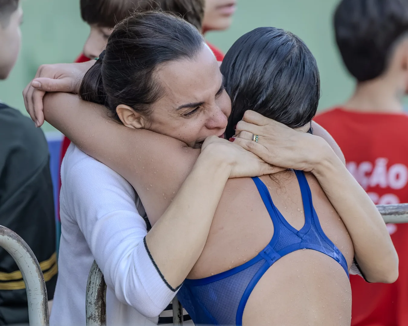 Adulta de blusa branca abraça uma nadadora de maiô azul à beira da piscina, olhos fechados, em gesto de acolhimento fora da água.
