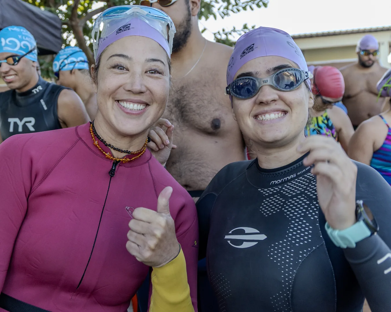 Duas nadadoras de touca lilás fazem sinal de positivo, uma de neoprene rosa e outra de preto