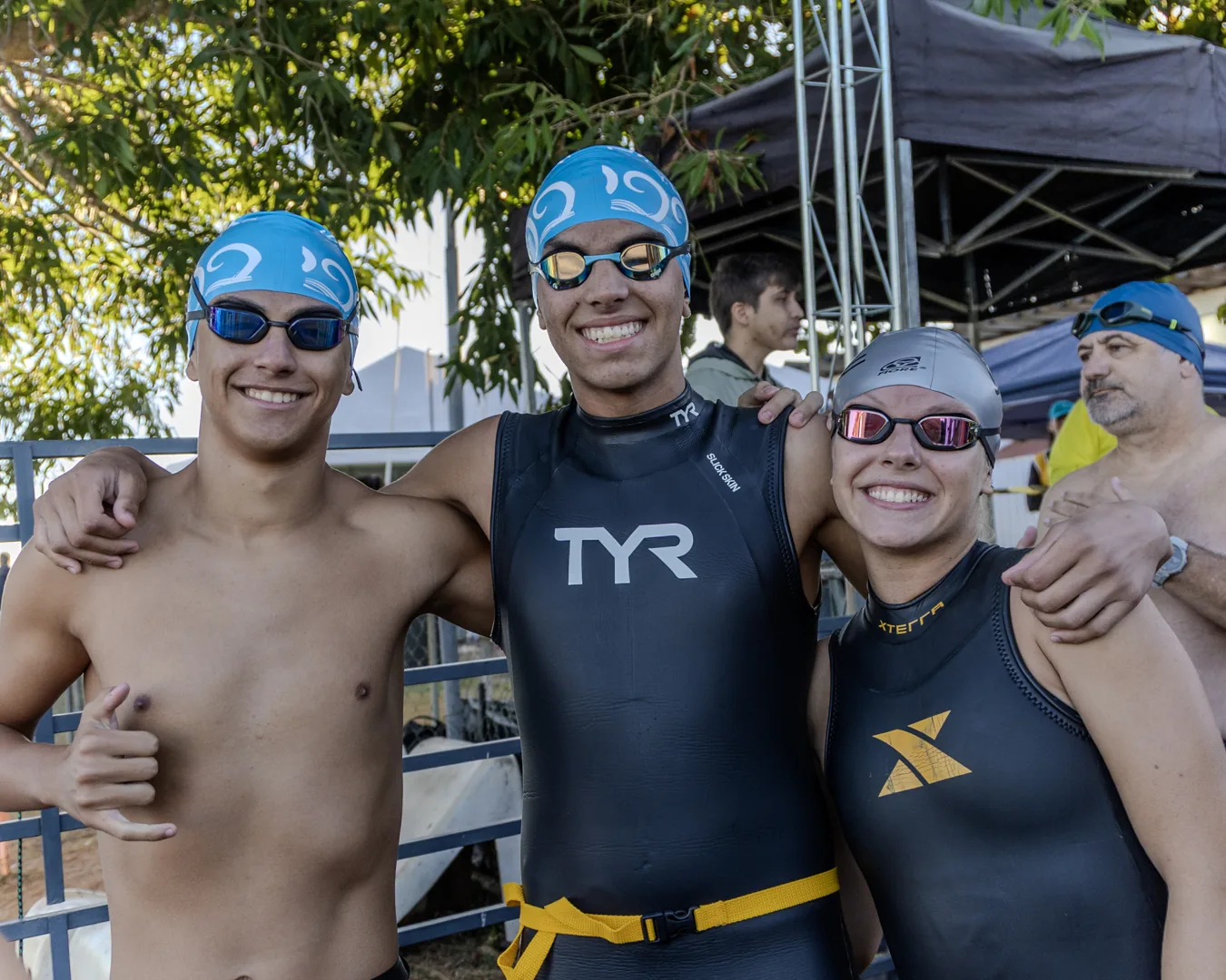 Três nadadores de touca azul e neoprene, abraçados pelos ombros, sorriem