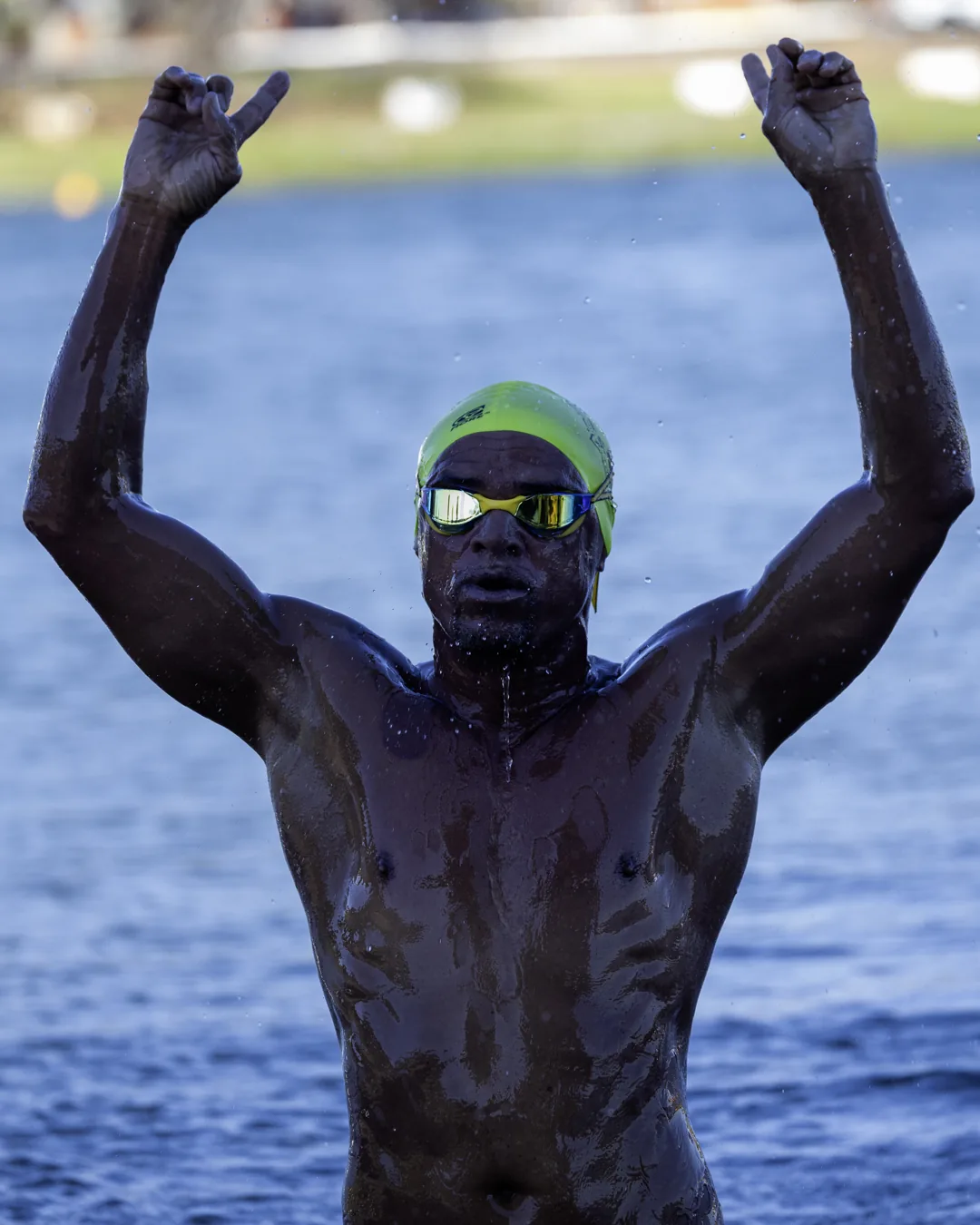 Nadador de touca amarela ergue os dois braços ao sair do lago