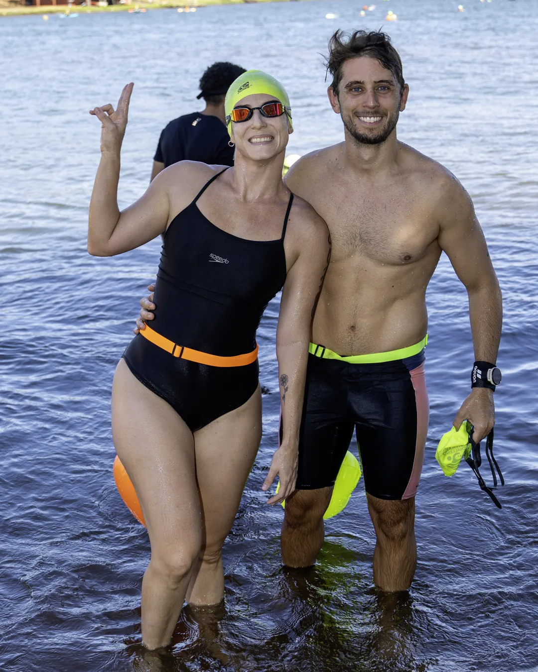 Nadadora e nadador saem do lago lado a lado, sorrindo, de touca amarela