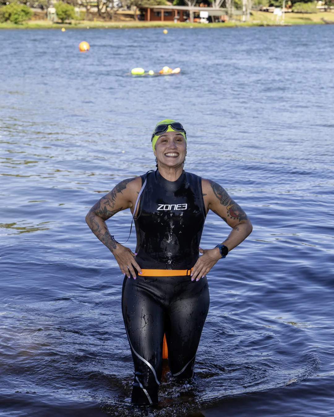 Nadadora de neoprene, mãos na cintura, sorri com o lago ao fundo