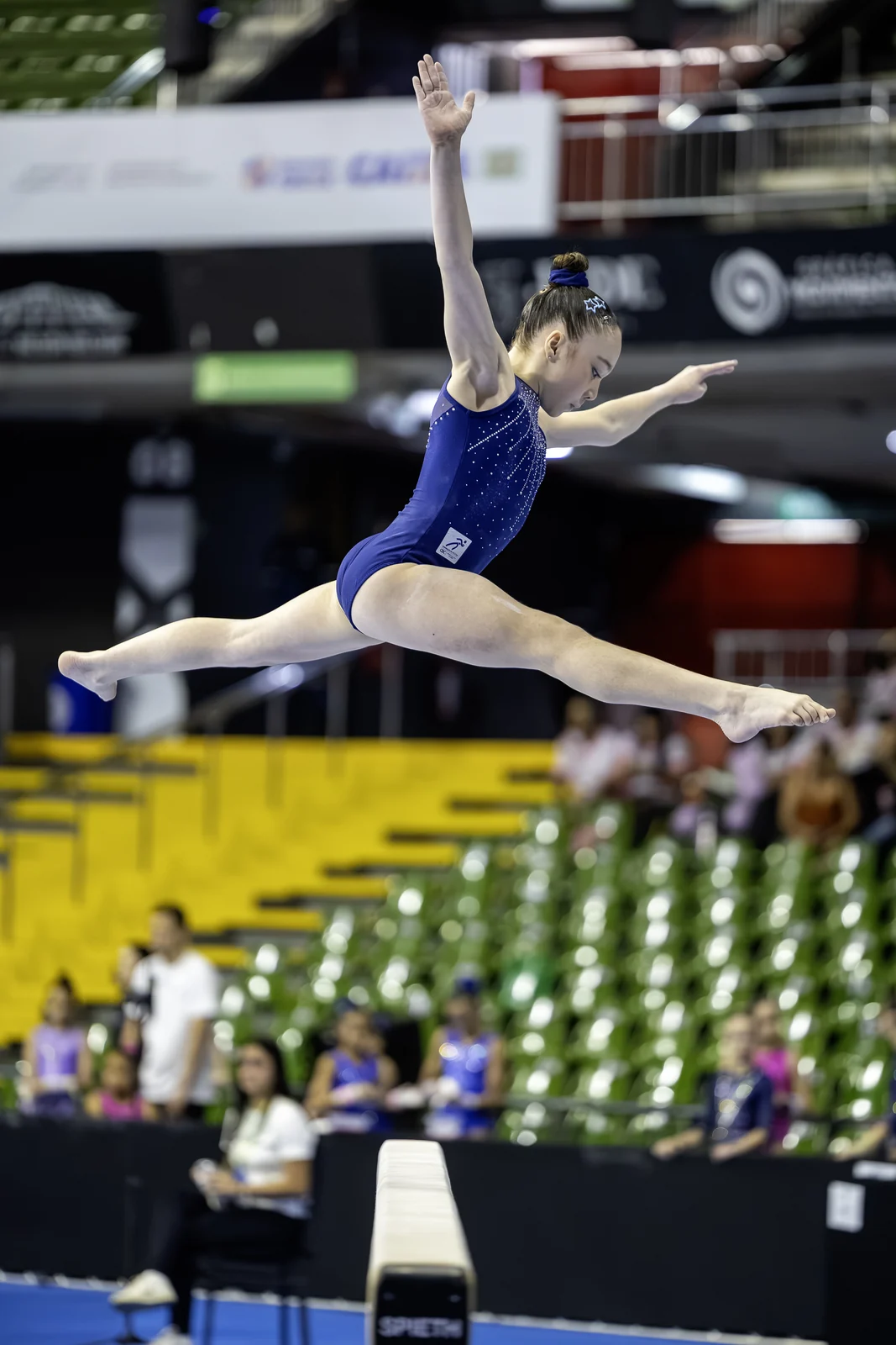 Ginasta em salto com as pernas abertas acima da trave, um braço estendido para cima