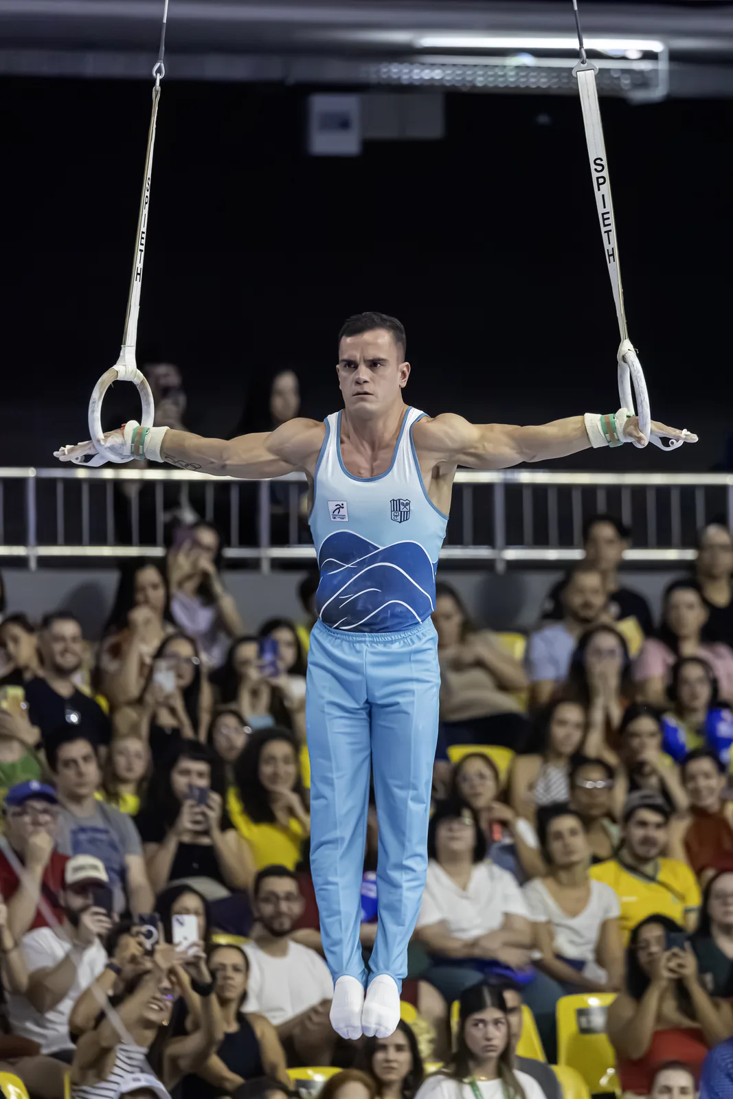 Ginasta sustentando o crucifixo nas argolas, diante do público