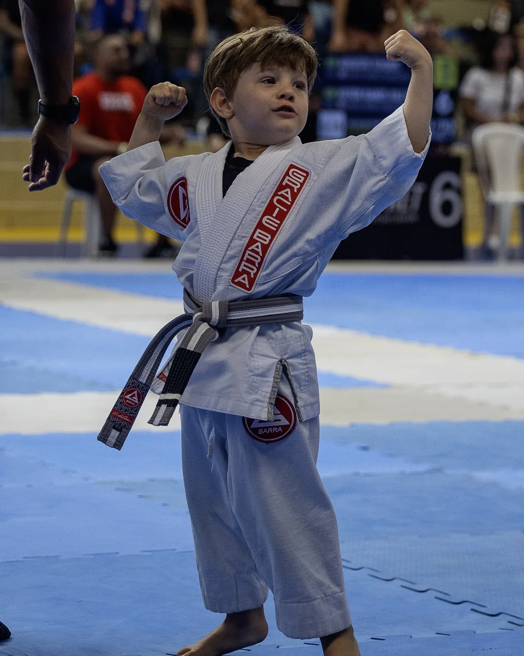 Menino de kimono branco e faixa cinza-branca flexiona os braços no tatame