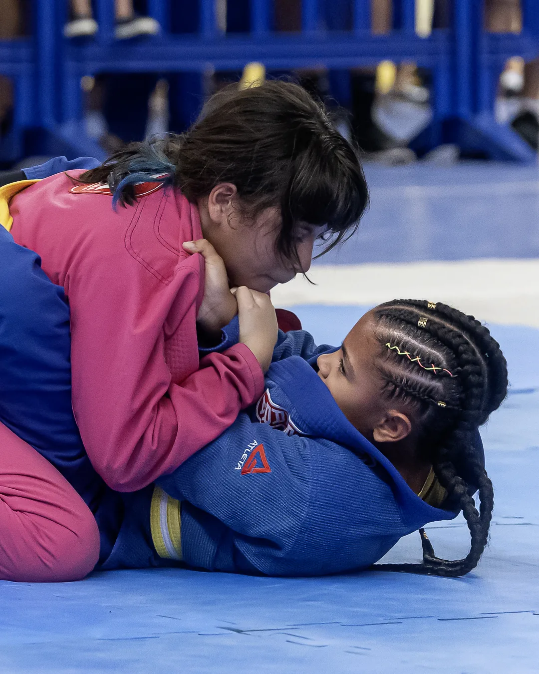Duas meninas em luta no solo, de kimono rosa e azul, rosto a rosto