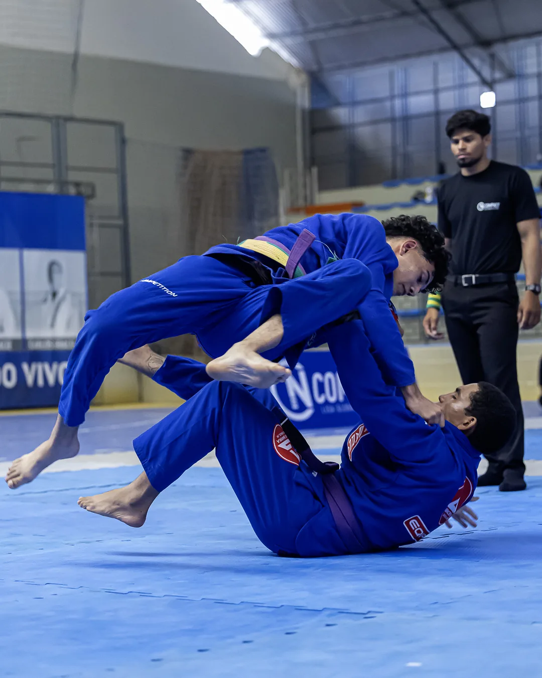 Atleta de kimono azul levanta a perna do adversário na raspagem, árbitro ao fundo