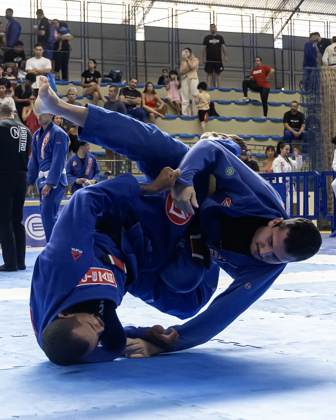Atleta de kimono azul invertido durante a luta, com a arquibancada ao fundo