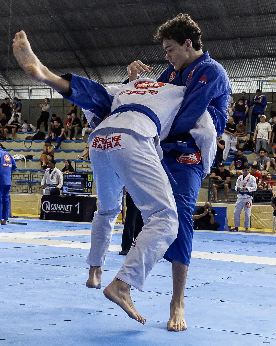 Atleta de kimono branco projetado no ar pelo adversário de azul, diante do painel da área 1