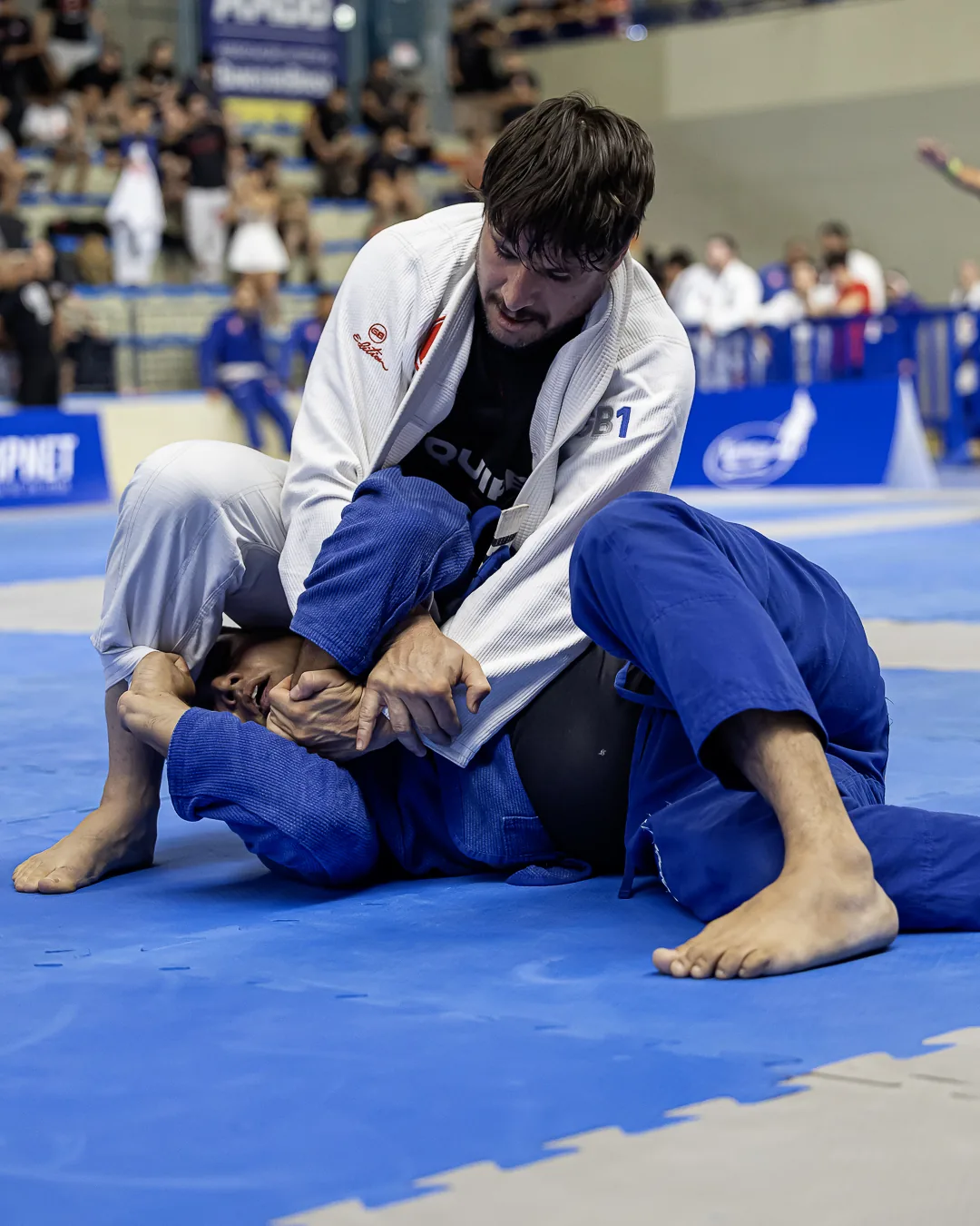 Atleta de kimono branco por cima, controlando o adversário de azul no solo