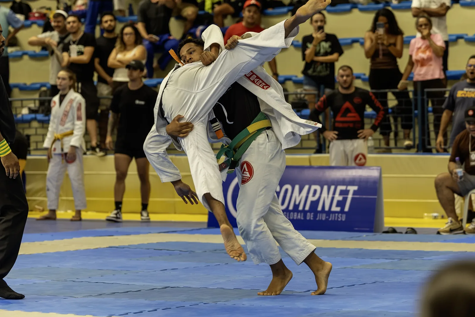 Atleta de kimono branco projeta o adversário no tatame, torcida ao fundo