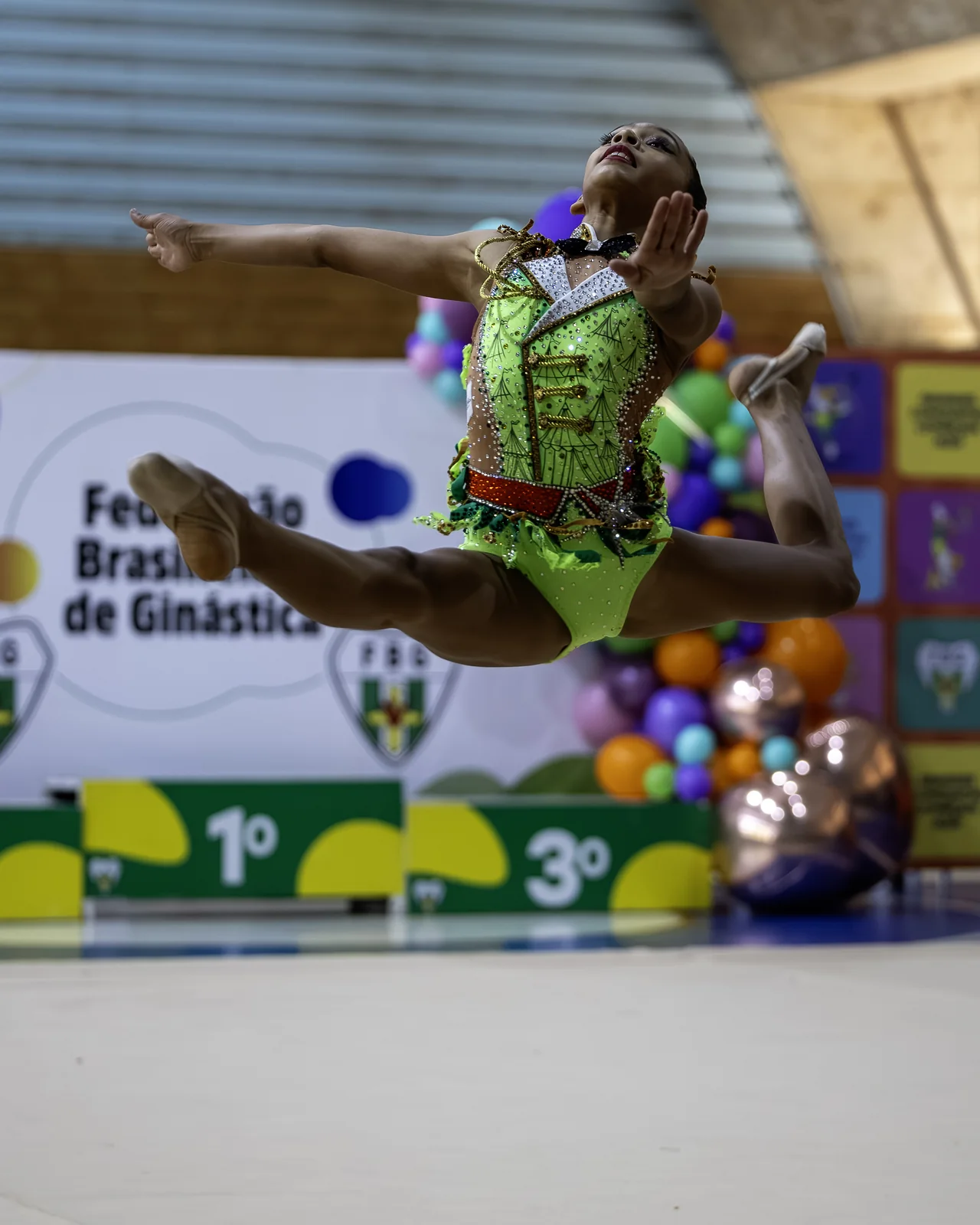 Ginasta em salto em split, com o pódio da Federação Brasiliense de Ginástica ao fundo