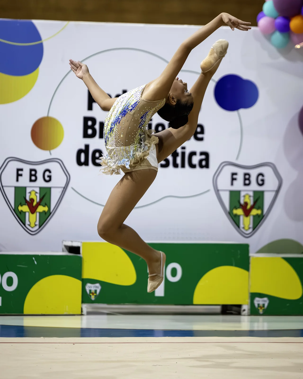 Ginasta em salto diante do painel da Federação Brasiliense de Ginástica