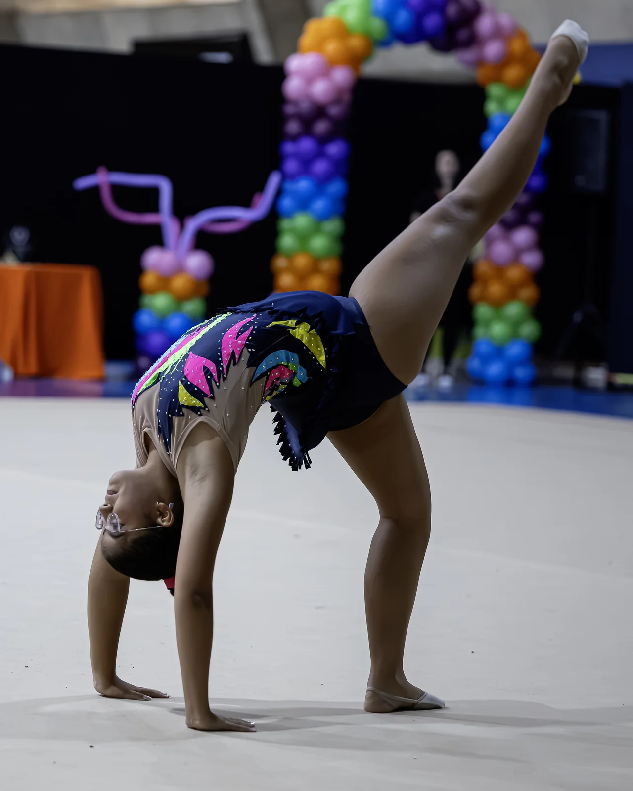 Ginasta em ponte com uma perna erguida na vertical, diante do arco de balões
