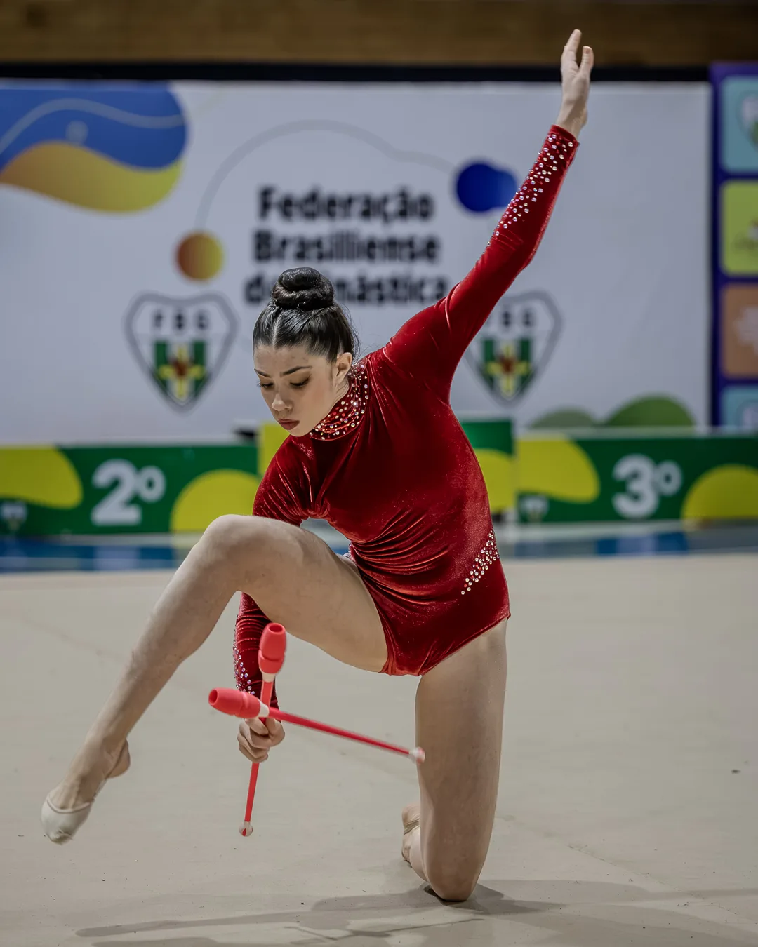 Ginasta em pose baixa com as maças diante do backdrop da Federação Brasiliense de Ginástica