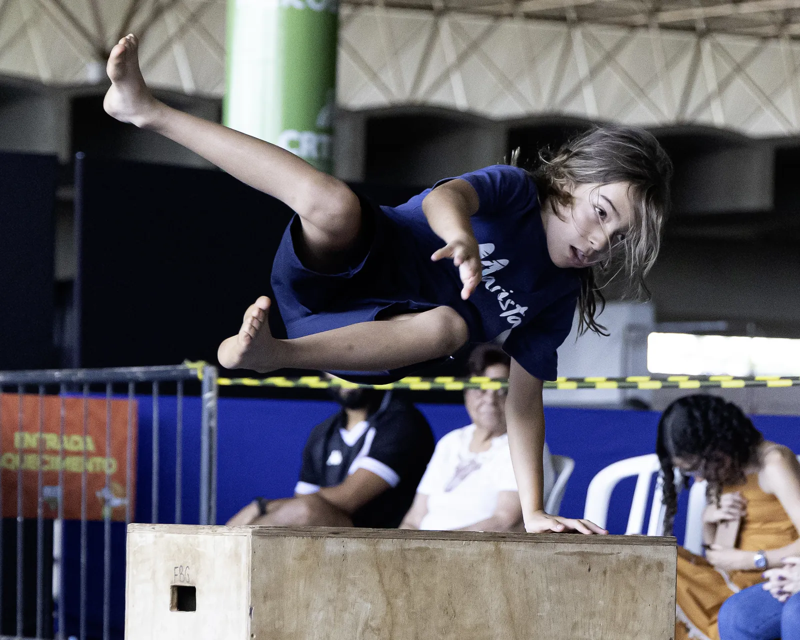 Menina de camiseta azul passa por cima de um caixote de madeira apoiada em uma das mãos, com as pernas estendidas para o lado