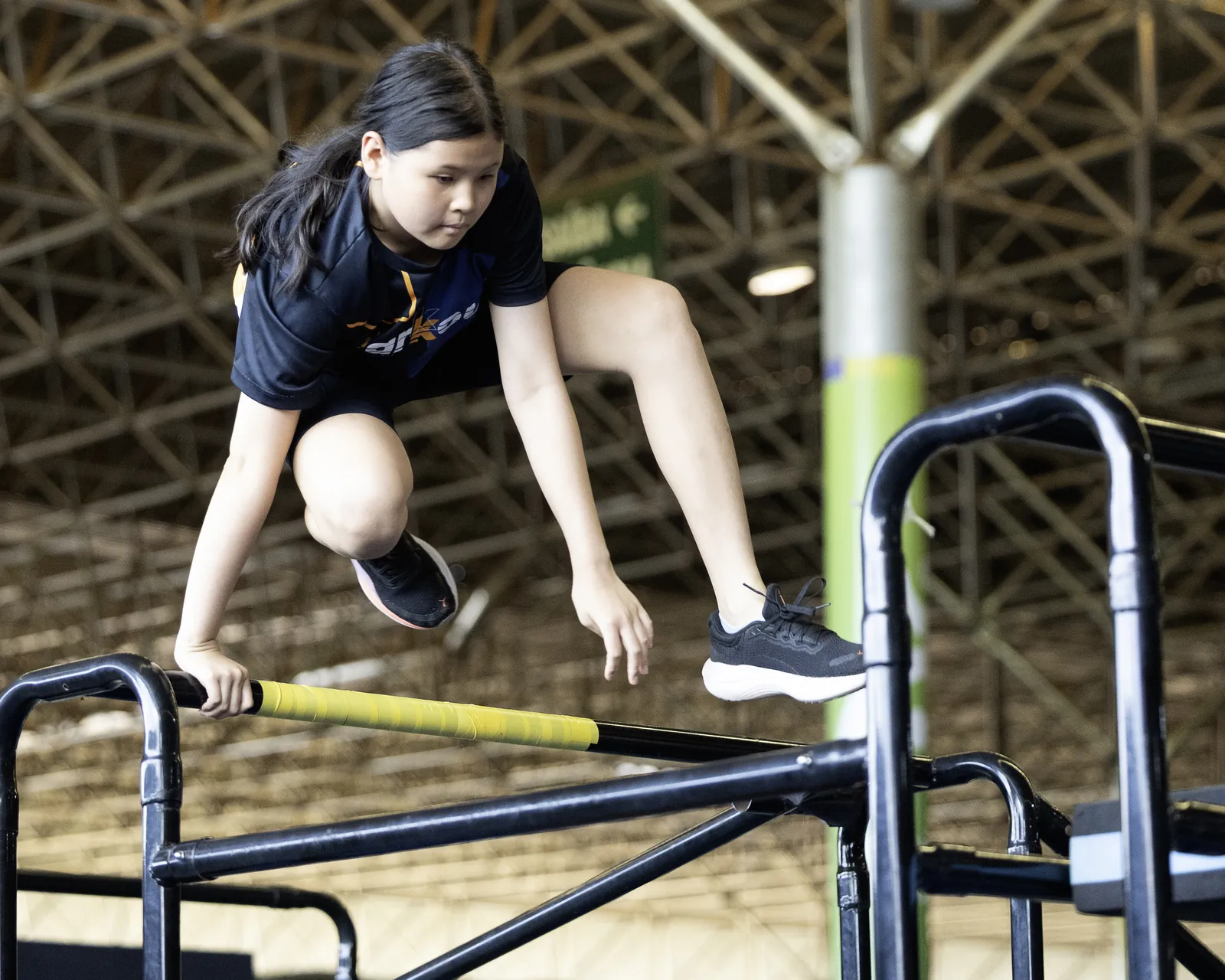 Menina de camiseta preta salta sobre uma barra metálica com fita amarela, sob a treliça do teto do pavilhão