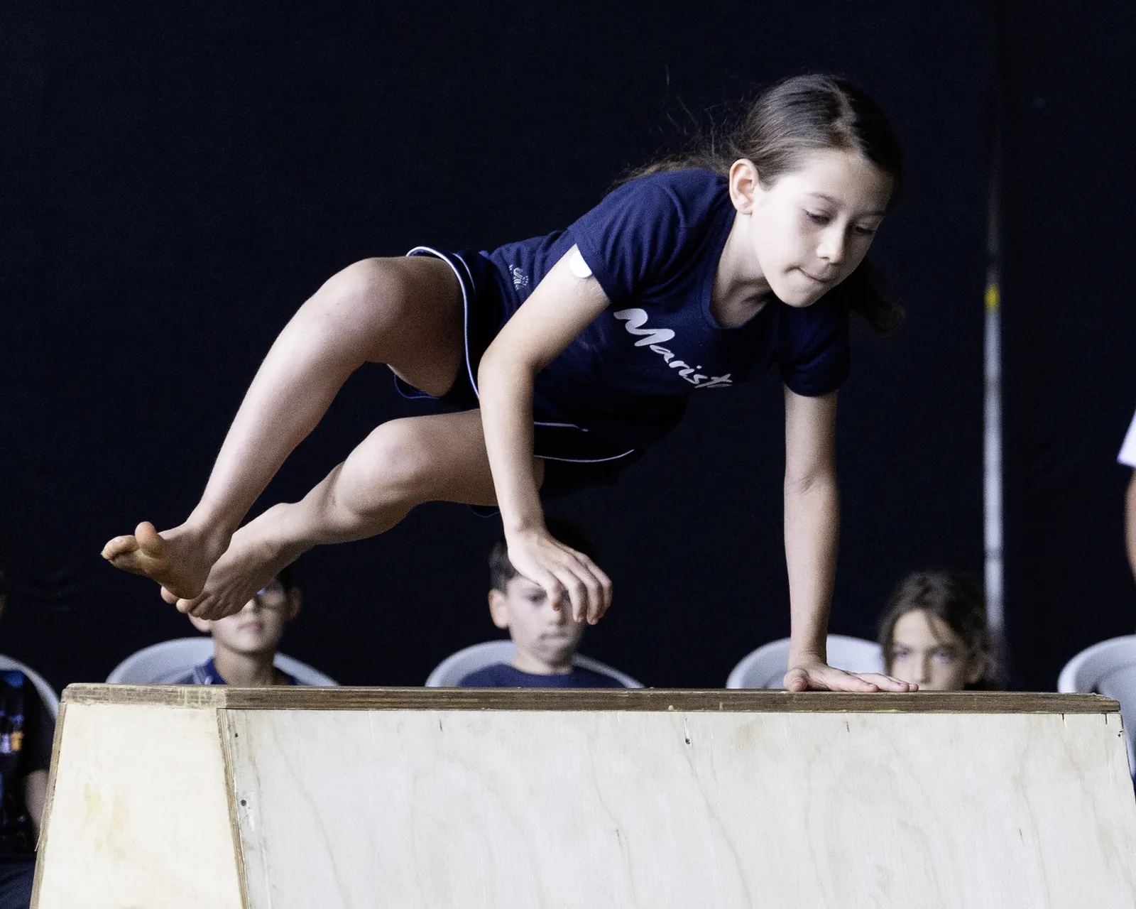 Menina de camiseta azul apoia as mãos no caixote de madeira com as pernas recolhidas no ar, diante de um fundo escuro
