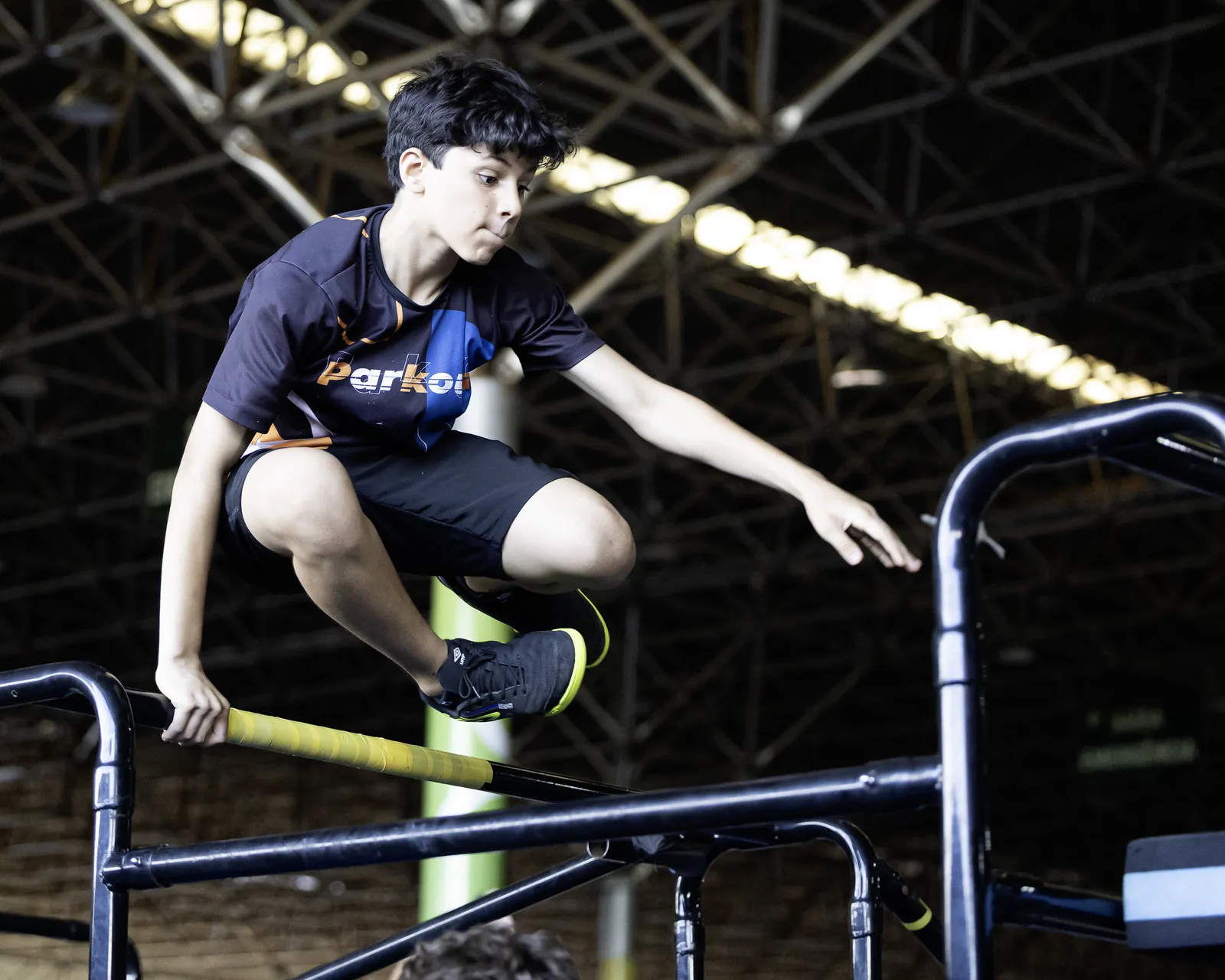 Garoto de camiseta azul e preta passa por cima de uma barra metálica, com o teto do pavilhão ao fundo