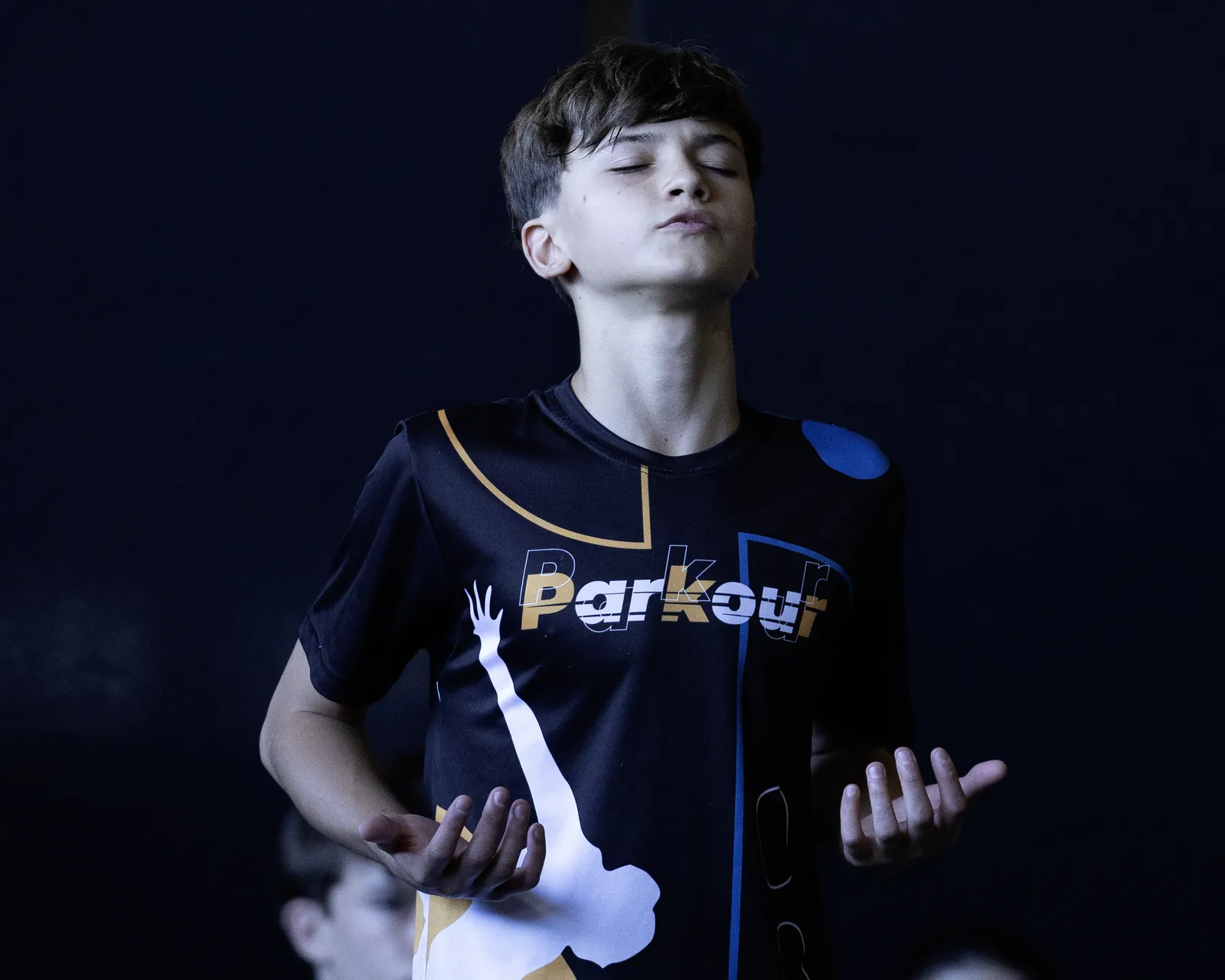 Garoto de camiseta com a palavra Parkour, de olhos fechados e mãos abertas, diante de um fundo escuro