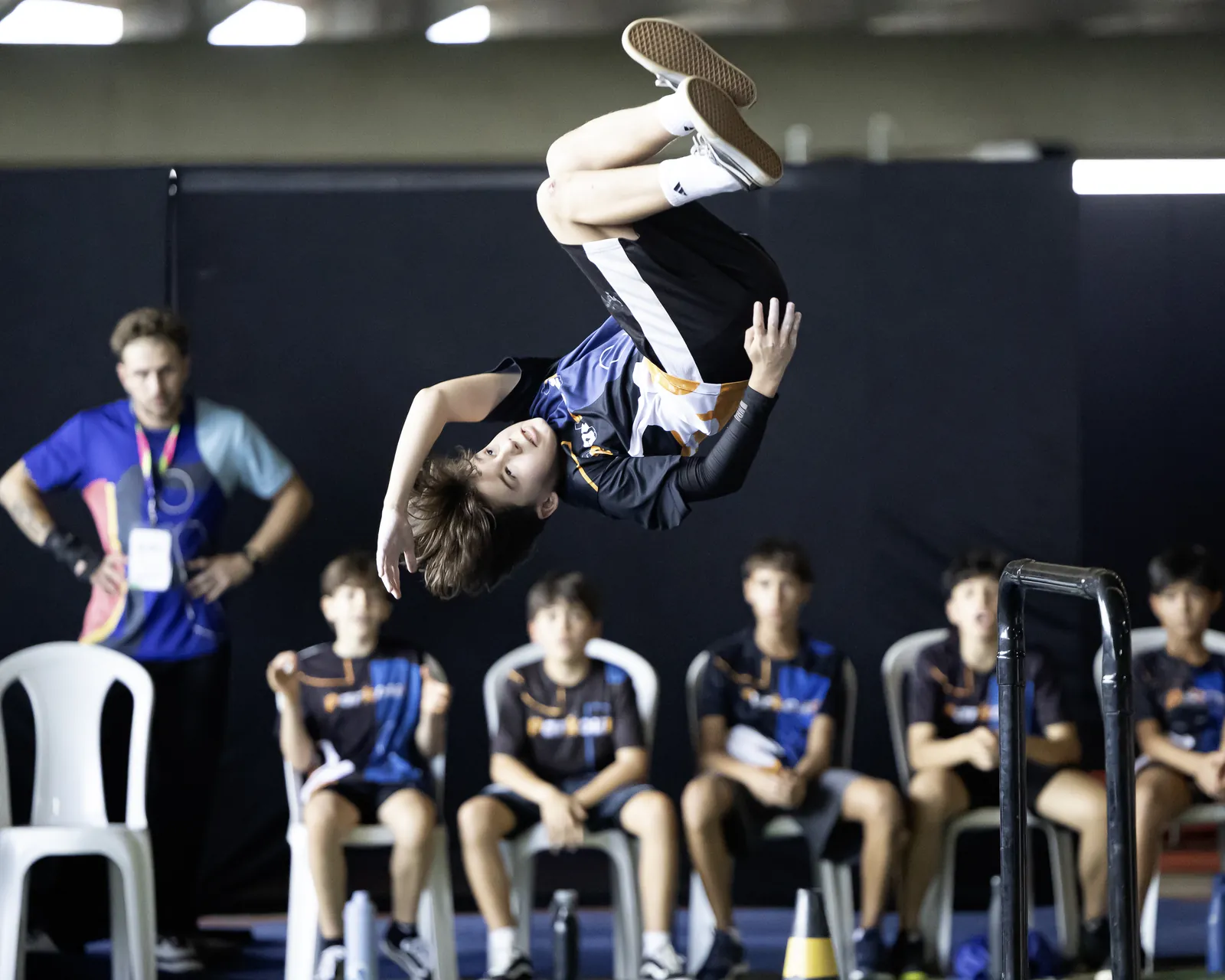 Atleta gira de cabeça para baixo no ar enquanto outros atletas, sentados em cadeiras, assistem ao fundo