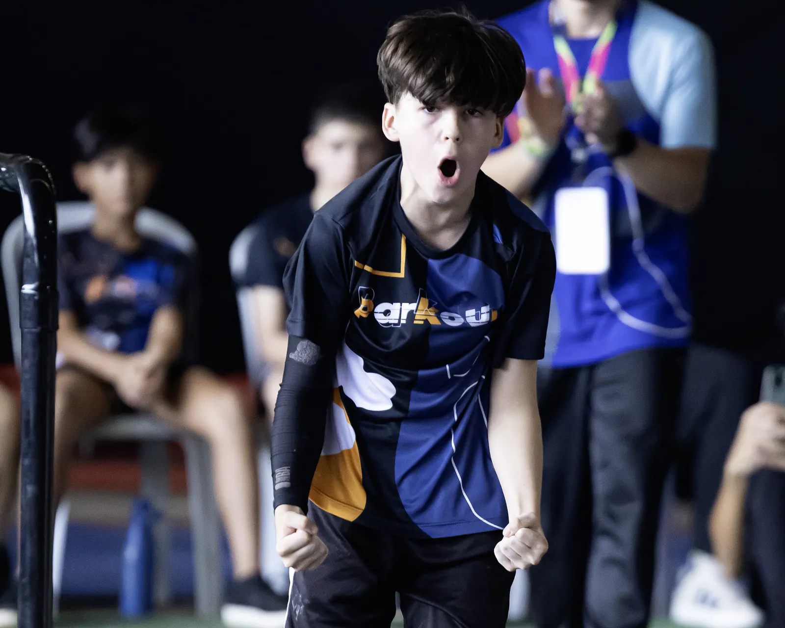 Garoto de camiseta Parkour comemora com os punhos fechados e a boca aberta, com atletas e plateia ao fundo
