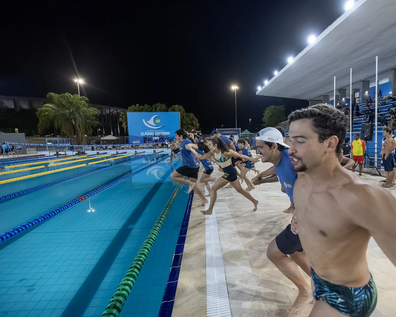 Atletas correm à beira da piscina iluminada, à noite, no fim da última etapa