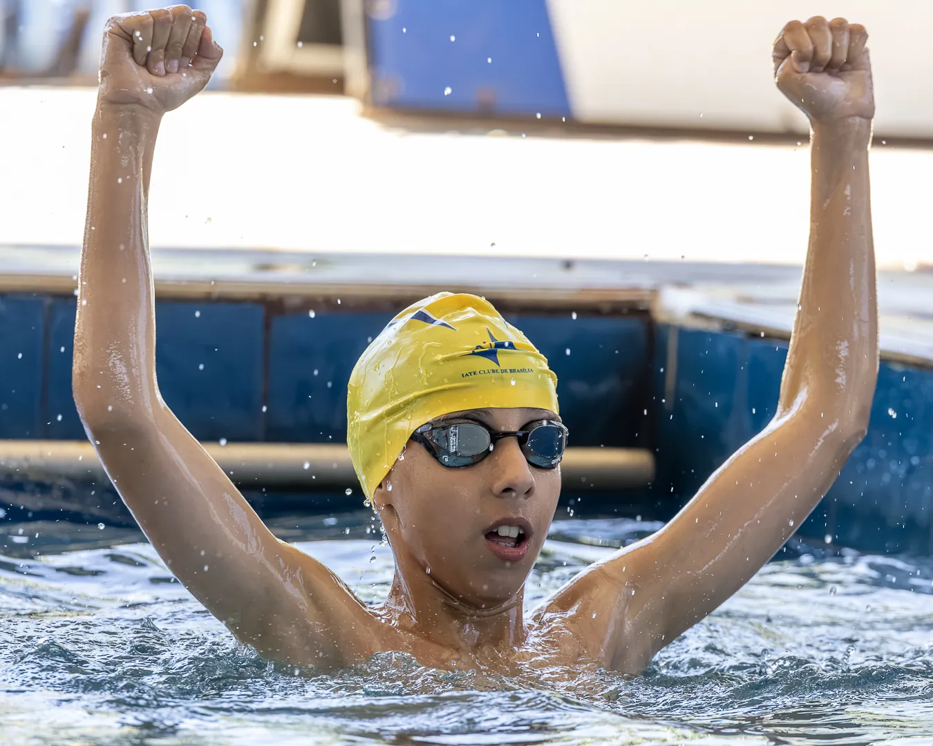 Nadador de touca amarela ergue os dois braços com os punhos fechados dentro da piscina