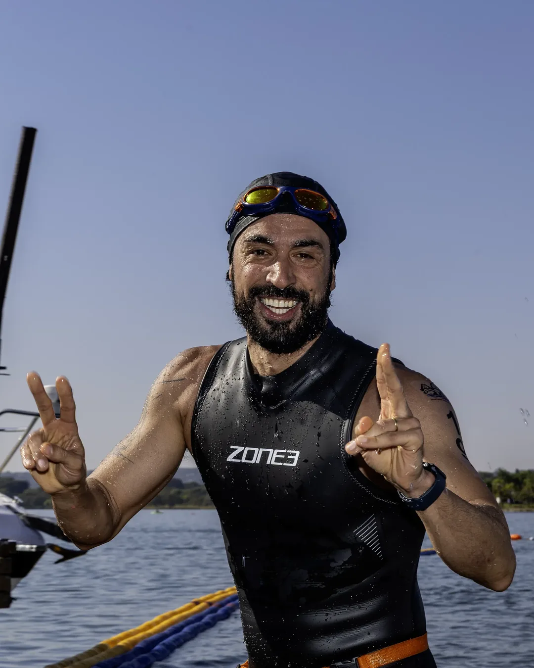 Nadador sorrindo com as duas mãos fazendo sinal de vitória, ainda de roupa de neoprene