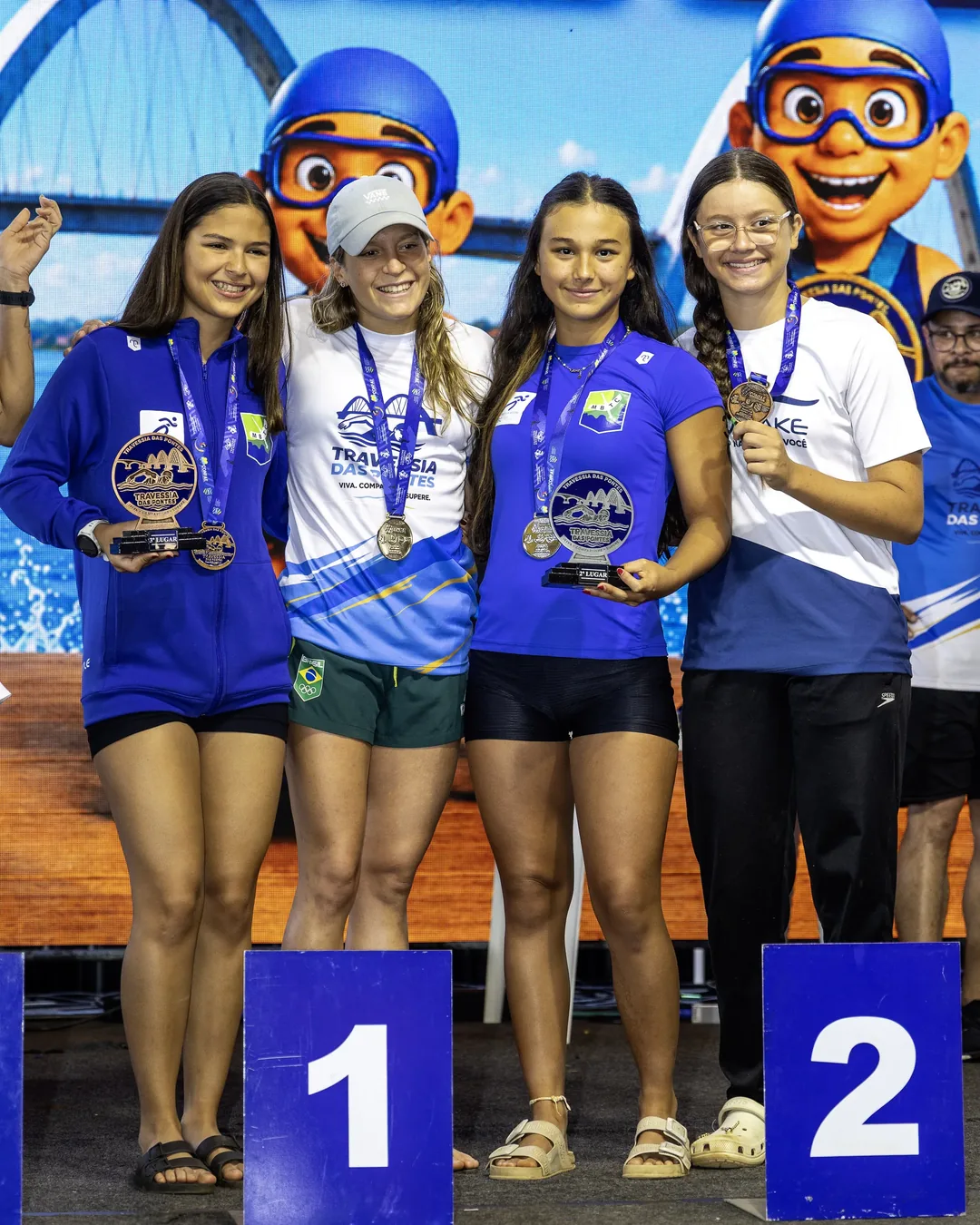 Quatro jovens nadadoras no pódio com troféus e medalhas, diante do painel do evento com a Ponte JK ilustrada