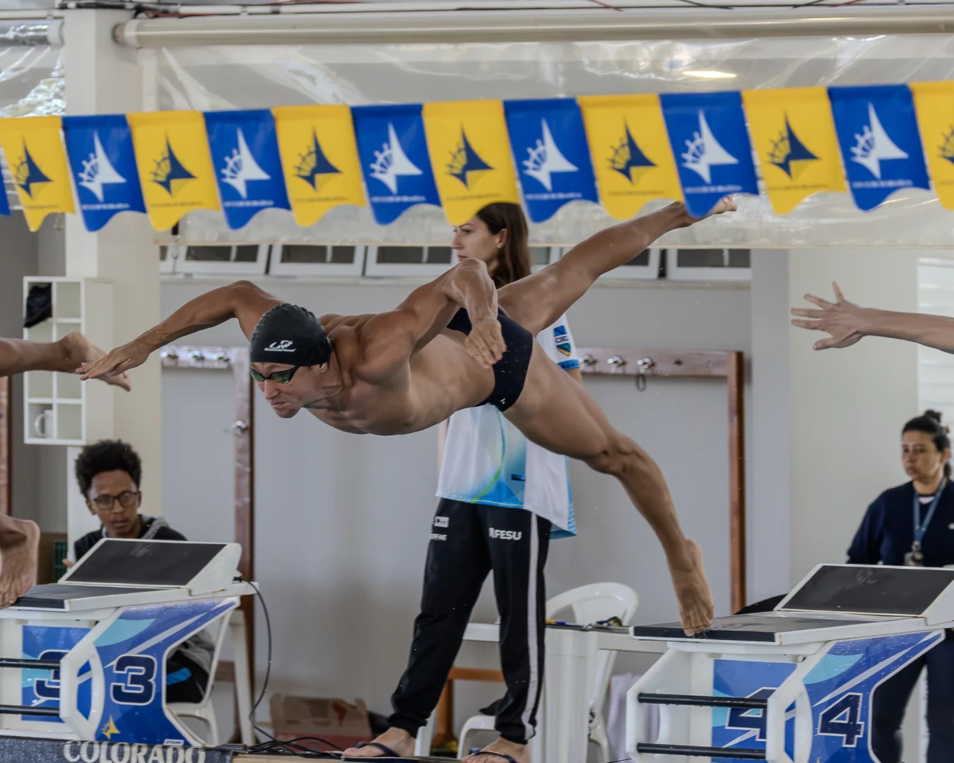 Nadador master saltando do bloco, corpo esticado no ar, sob as bandeirolas do clube